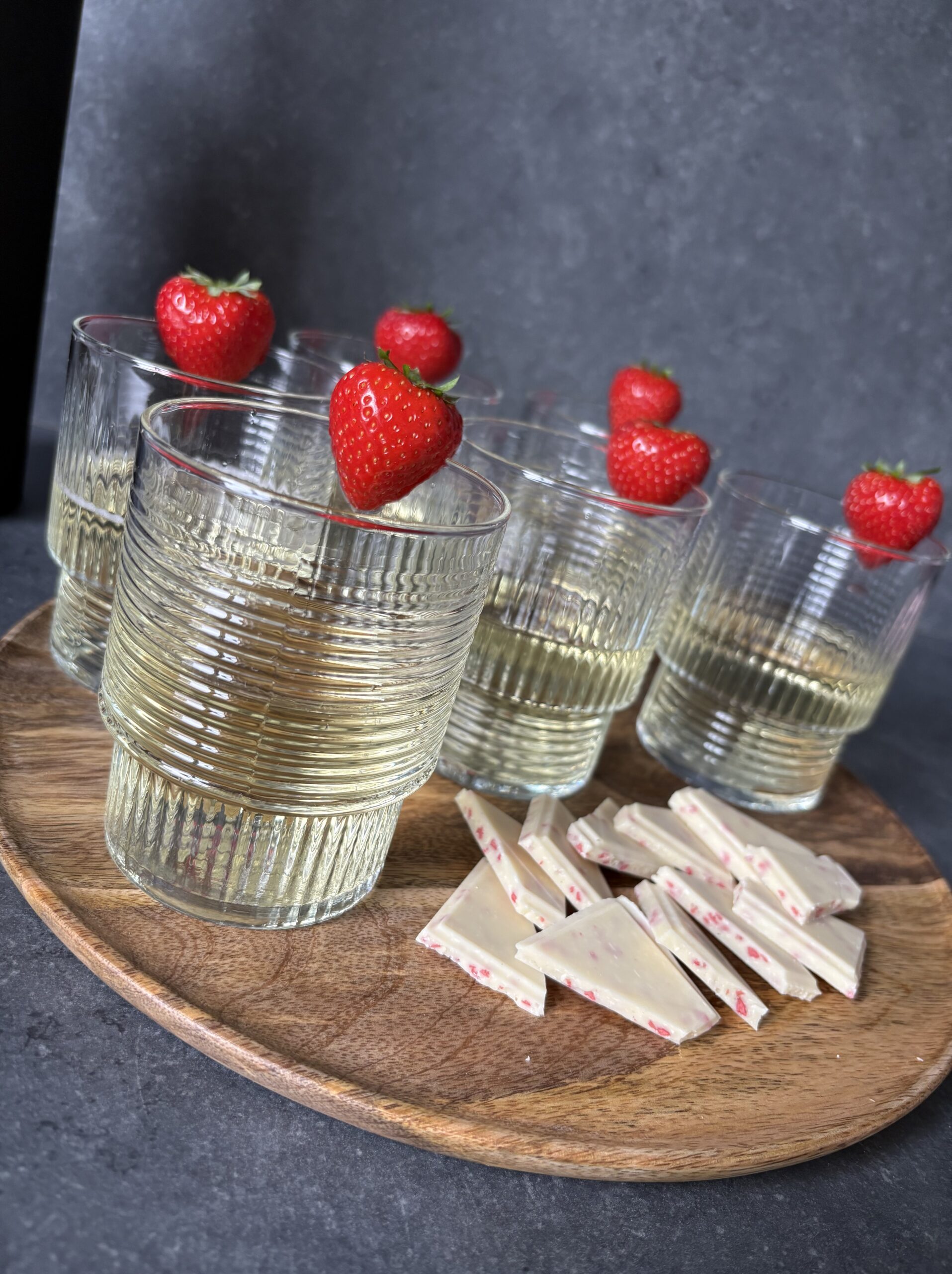 Glasses of white wine garnished with strawberries on a wooden tray next to pieces of strawberry white chocolate